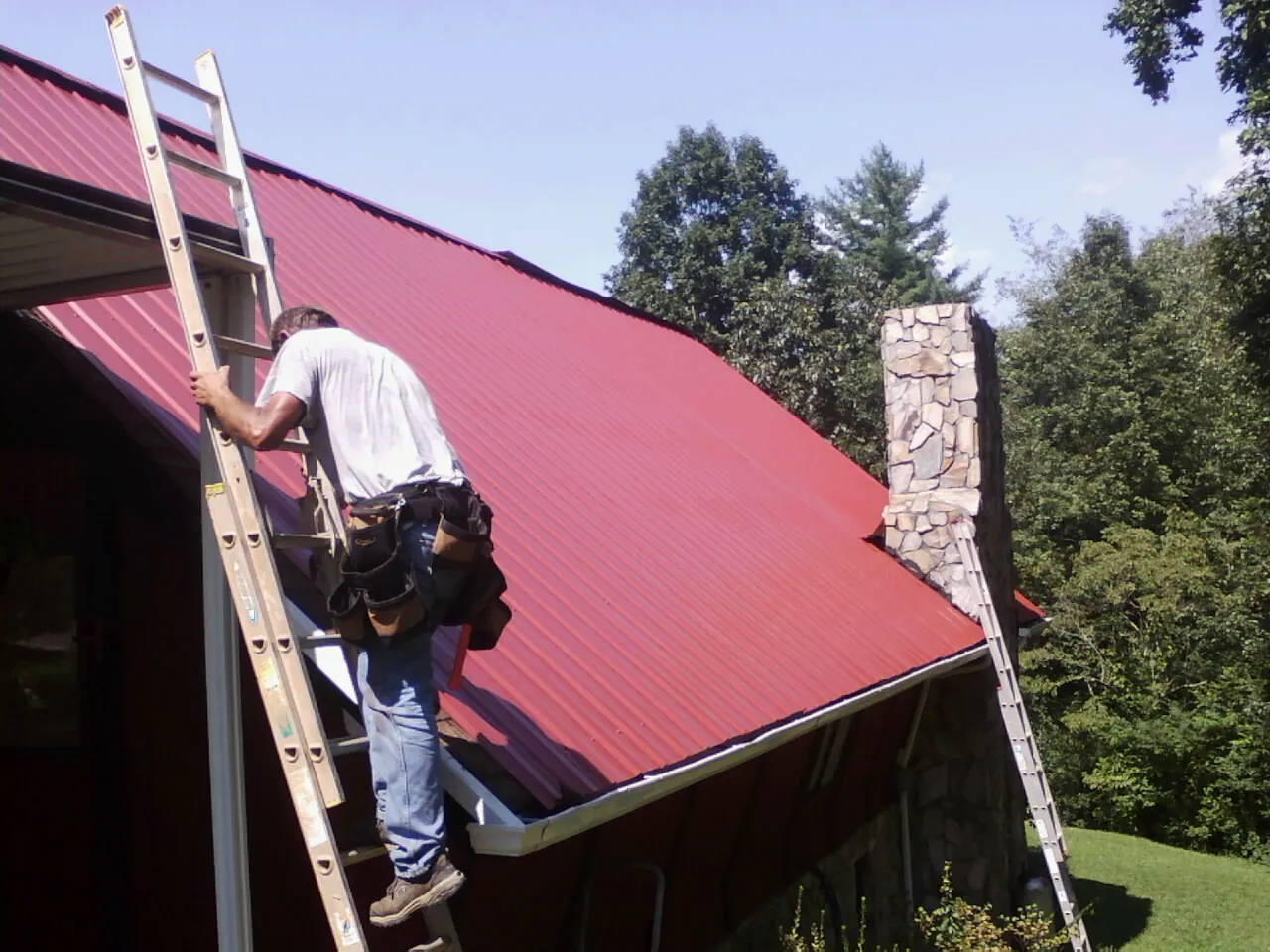 Roofer on a ladder beside a red metal roof and stone chimney — Soffit & Fascia Repair by Roofing Brothers in Thompsons